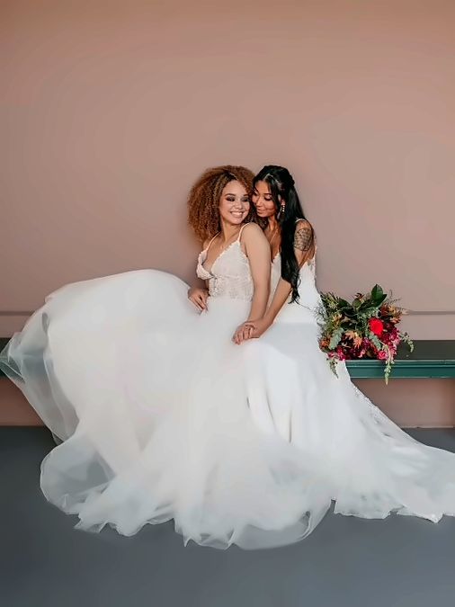 Two brides in white gowns embracing, soft glam makeup